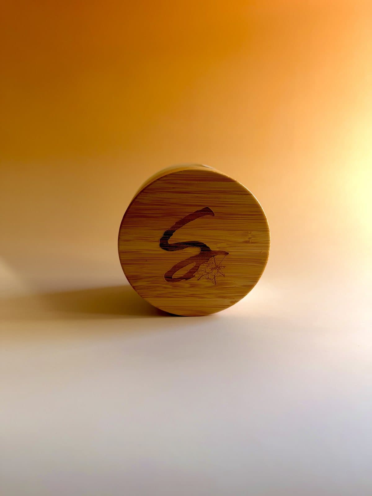A single bamboo lid engraved with the Scented Shea mark, resting on a warm amber ground